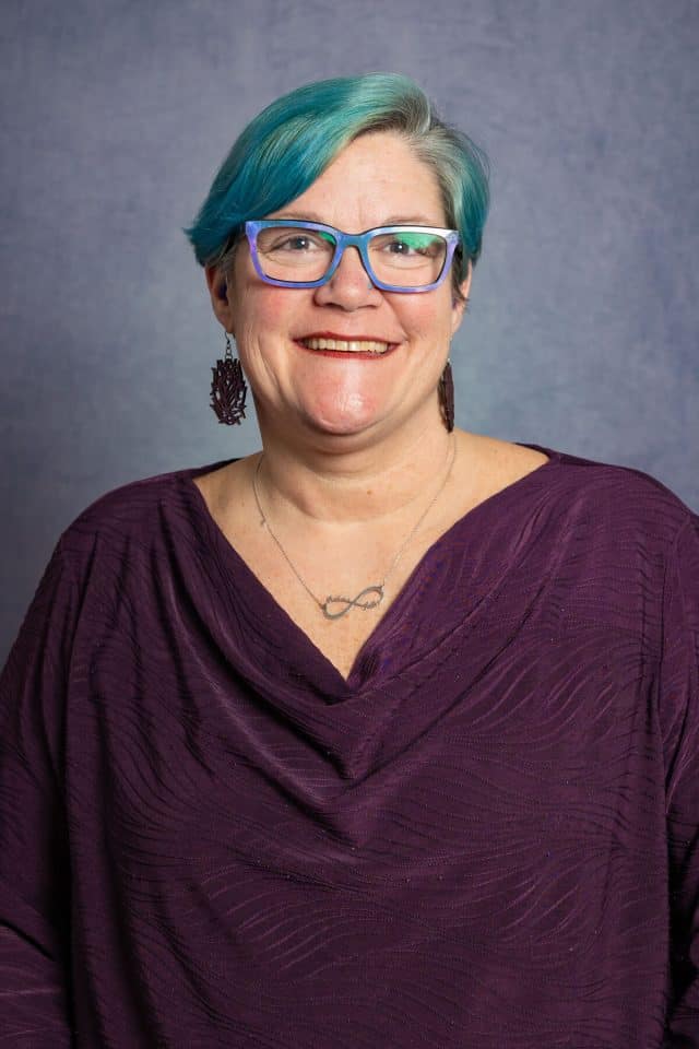 White woman with a teal-colored pixie cut wearing blue glasses, earrings and a purple blouse